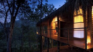 A wooden cabin with a thatched roof, elevated on stilts, and illuminated by interior lighting. - Guava House by The Serendipity Collection (Hemmathagama)