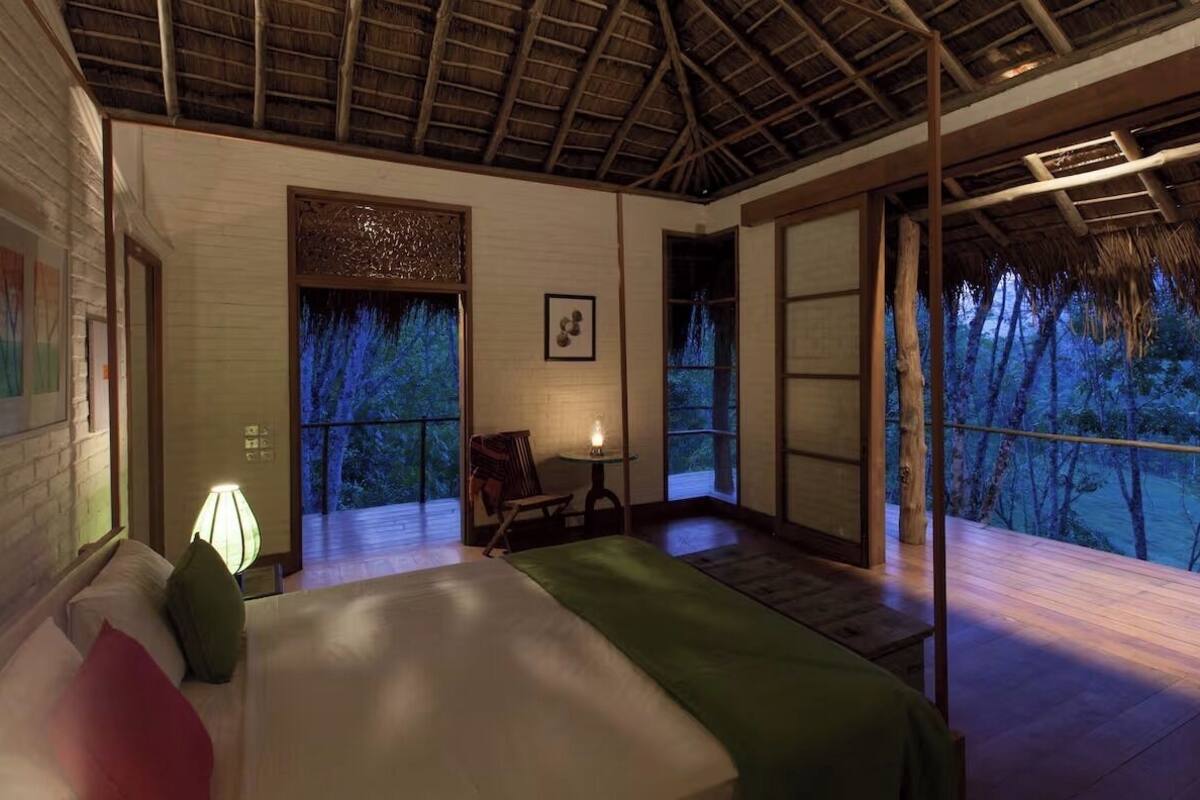 Un dormitorio con una cama, una mesita de noche, una lámpara y vistas a árboles a través de ventanas amplias.