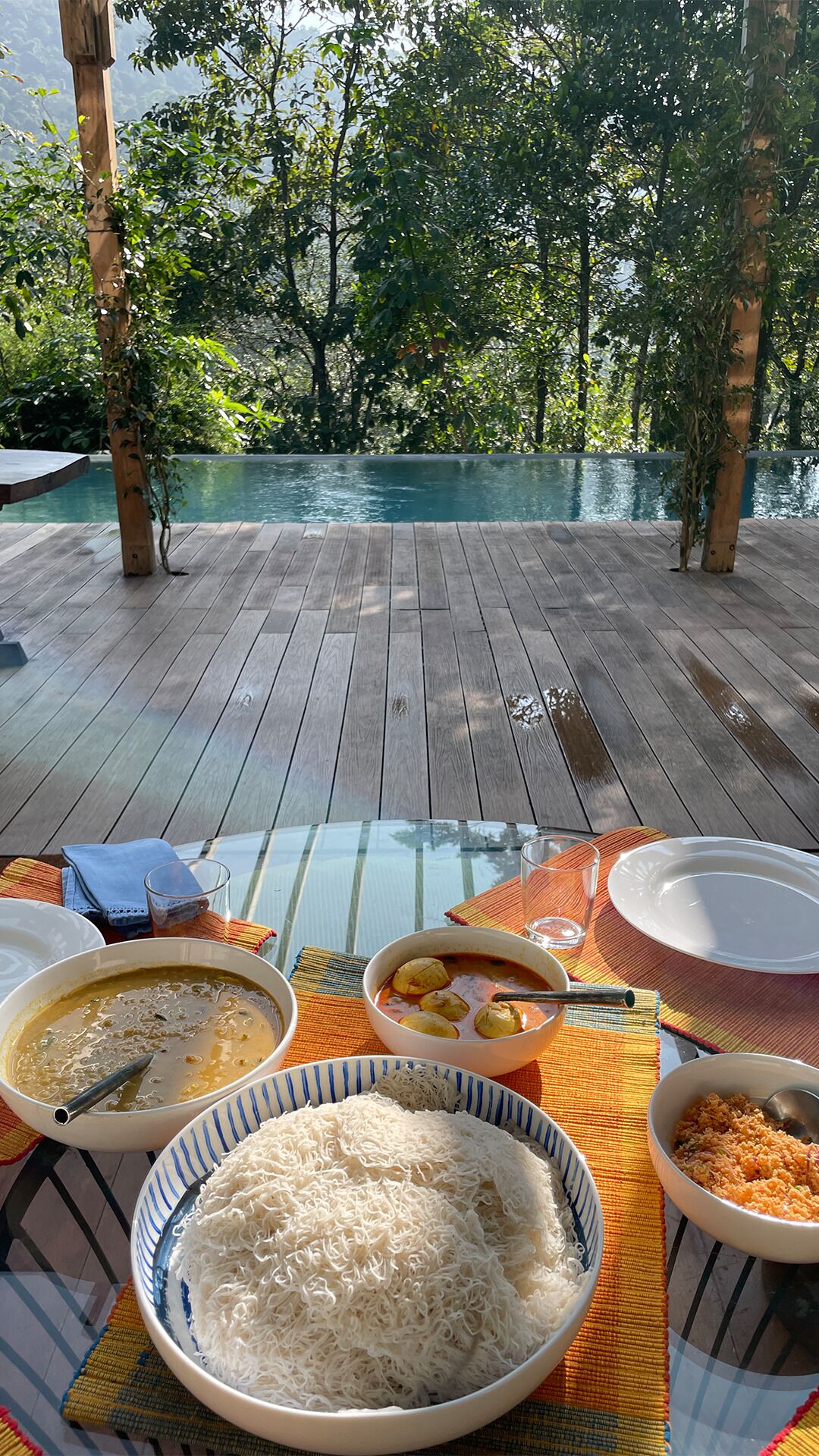 An outdoor dining area with a pool, wooden deck, and lush greenery in the background.