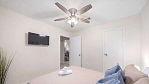 Una camera d'albergo con un letto, una ventola a soffitto, una TV a schermo piatto, una porta e una pianta in vaso.