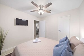 A hotel room with a bed, a ceiling fan, a flat-screen TV, a door, and a potted plant. - Beach Walk Home, Fenced Yard & Fire Pit (Seabrook)