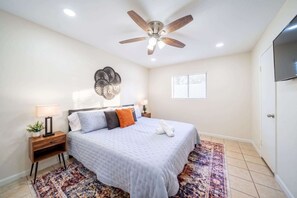 Une chambre à coucher comprenant un lit, un ventilateur de plafond, un élément décoratif fixé au mur, une table de chevet avec une lampe et une fenêtre avec des stores.