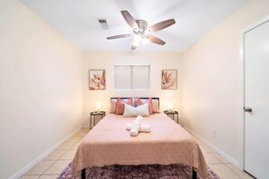 Un dormitorio con cama, ventilador de techo, dos mesitas de noche con lámparas, una ventana con persianas y dos cuadros colgados en la pared.