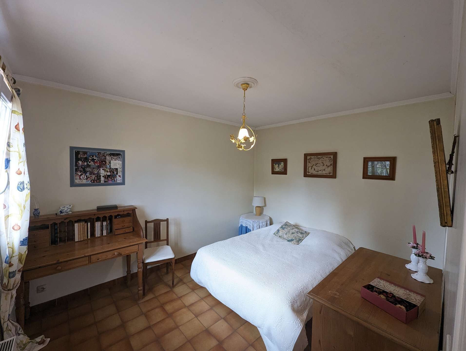 A bedroom with a bed, a desk, a chair, a small table with a vase, and a hanging light fixture.