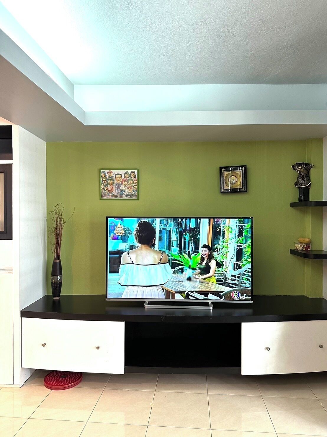 Sala de estar con una televisión de pantalla plana sobre un pedestal negro, paredes verdes y un mueble blanco debajo.