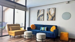 Ein modernes Wohnzimmer mit einem blauen Sofa, einem senffarbenen Sessel und zwei weißen Hocker. An der Wand befinden sich abstrakte Gemälde und ein runder Spiegel.