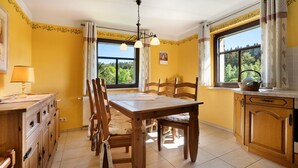 Una cocina con una mesa de madera, sillas, un candelabro colgante y ventanas grandes con cortinas.