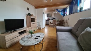 Ein Wohnzimmer mit Fernseher, Sofa, Esstisch und einer Küche im Hintergrund.
