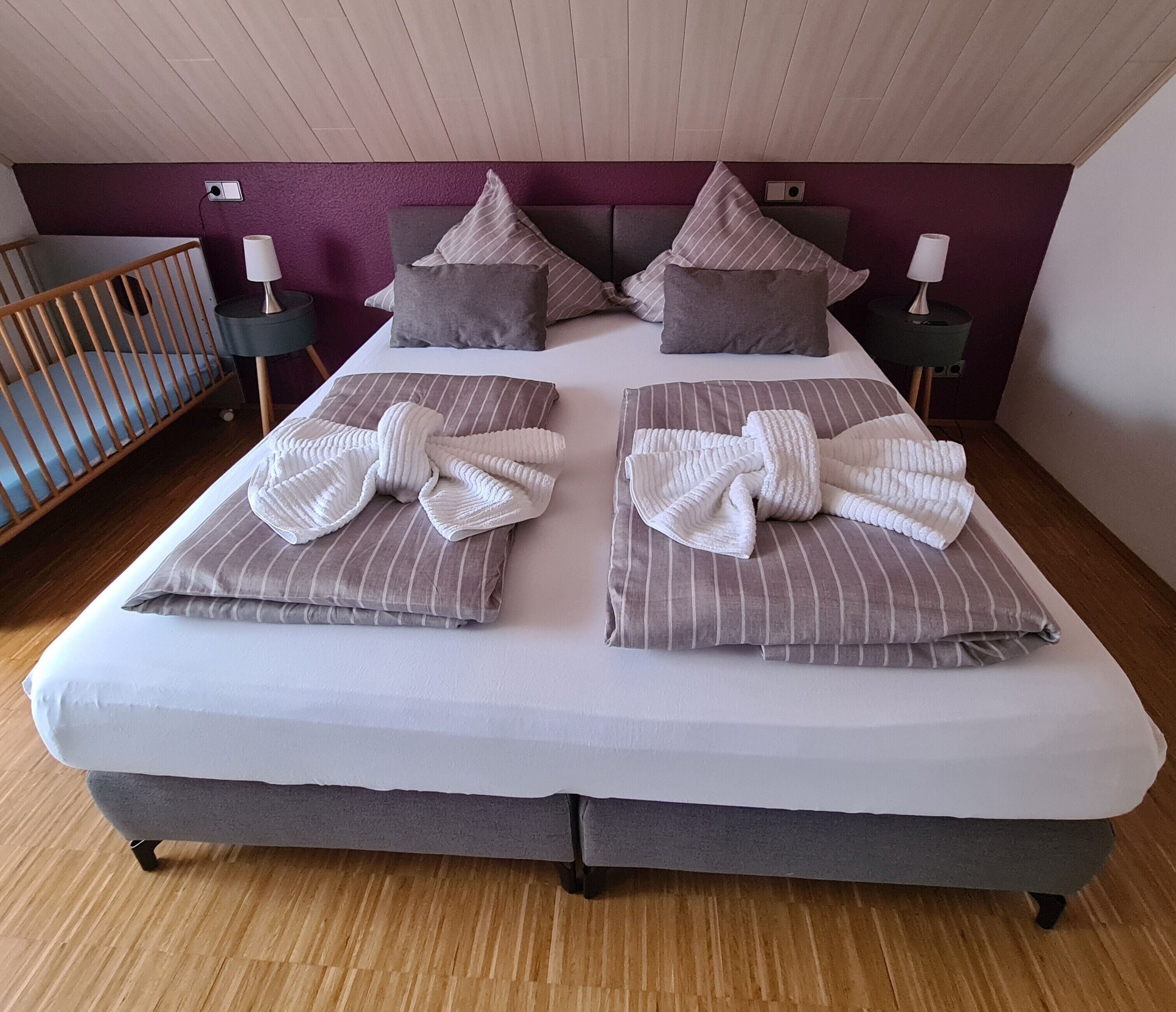 Ein Doppelbett mit gestreiften Kissen und einem gefalteten Handtuch, ein Holz-Babybett und eine violette Akzentwand.