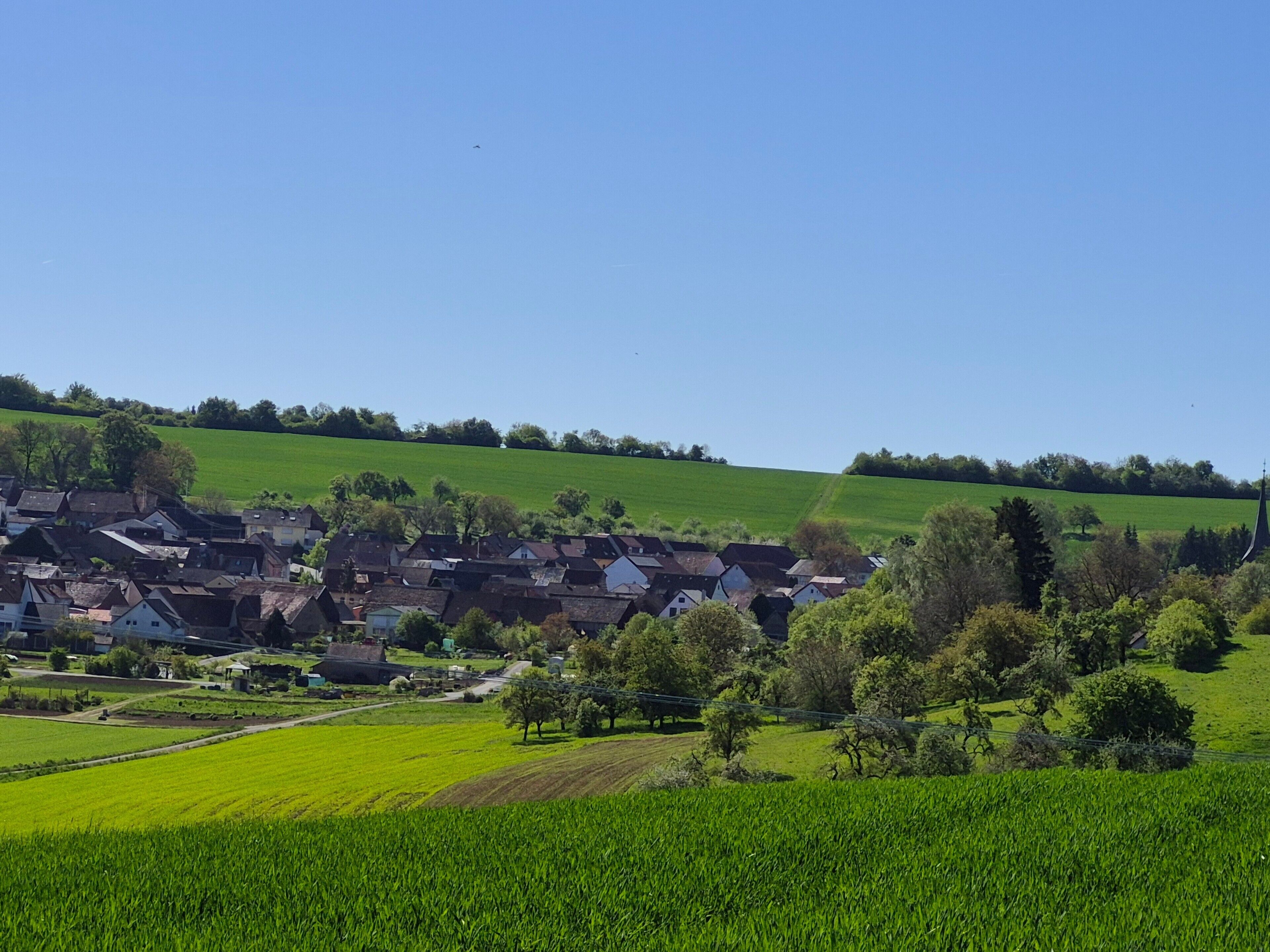 Un pueblo rural con casas tradicionales, campos verdes y la aguja de una iglesia.