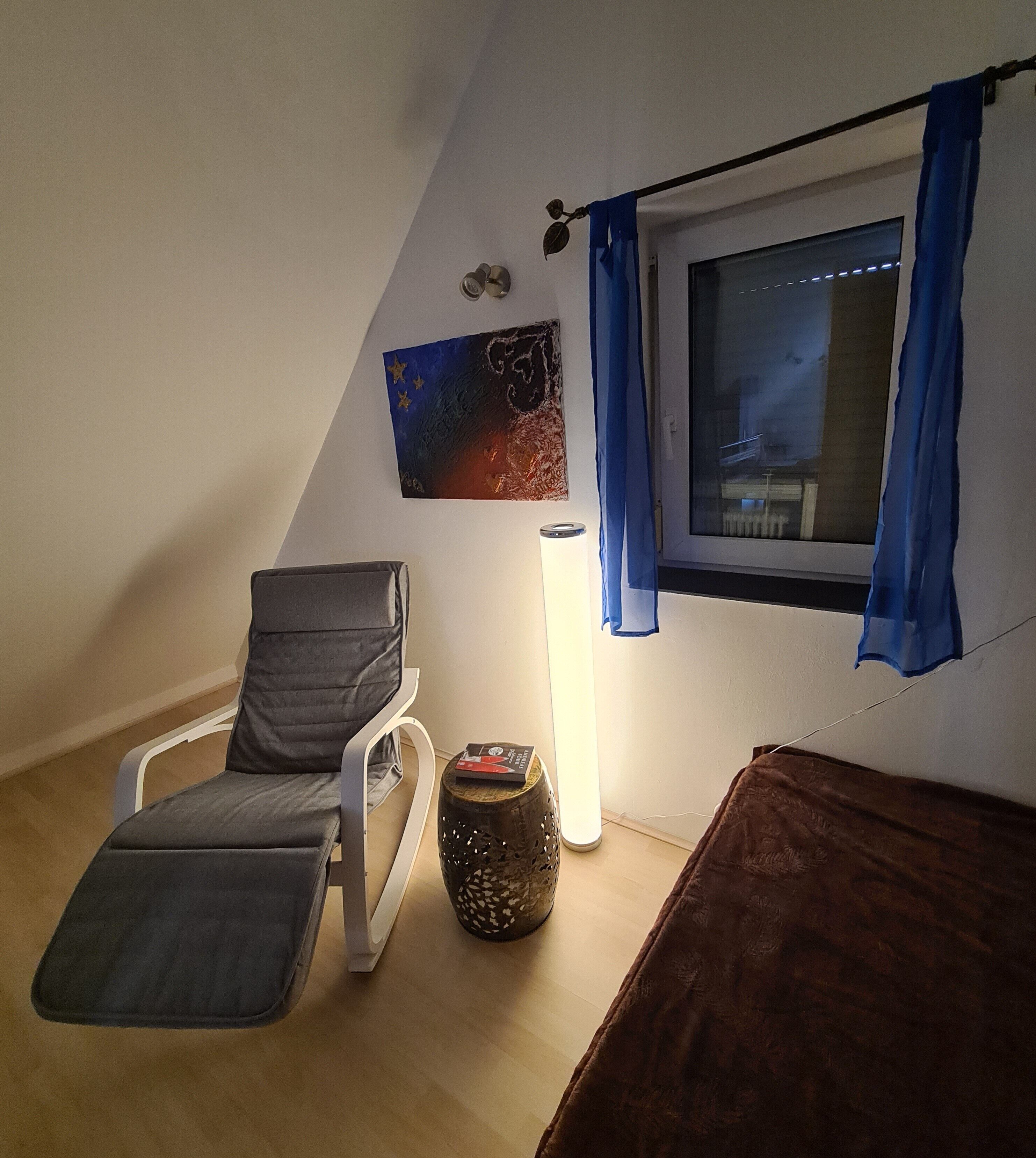 Una stanza con un letto, una sedia, una lampada, una finestra con tende e un quadro appeso al muro.