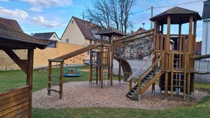 Ein Spielplatz mit Holzelementen, darunter eine Rutsche und ein Kletterbereich, umgeben von einer Rasenfläche und Wohnhäusern im Hintergrund.