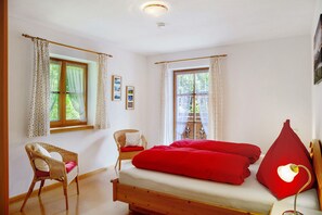 Una habitación con una cama, dos ventanas con cortinas, una silla de mimbre y una lámpara de pared.