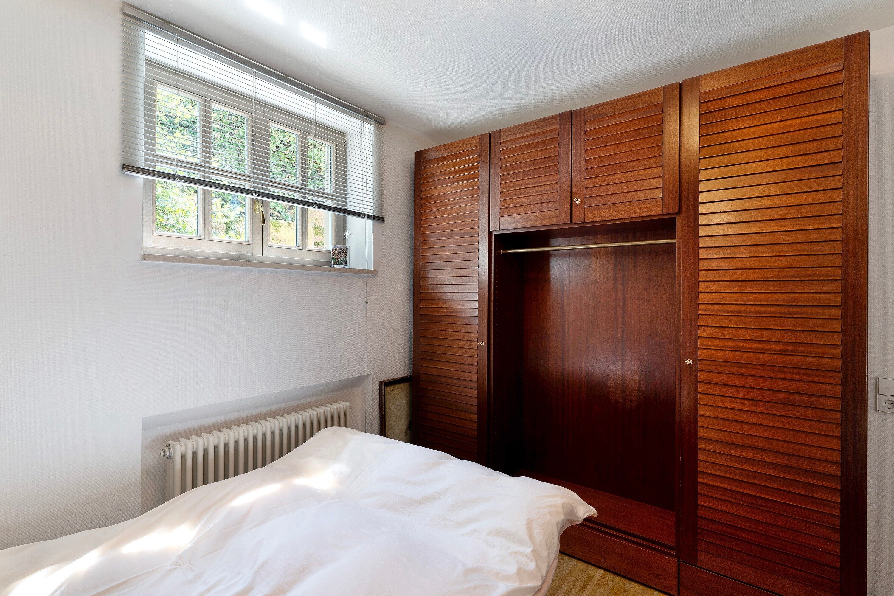A bedroom with a bed, a window with blinds, a radiator, and a wooden wardrobe.