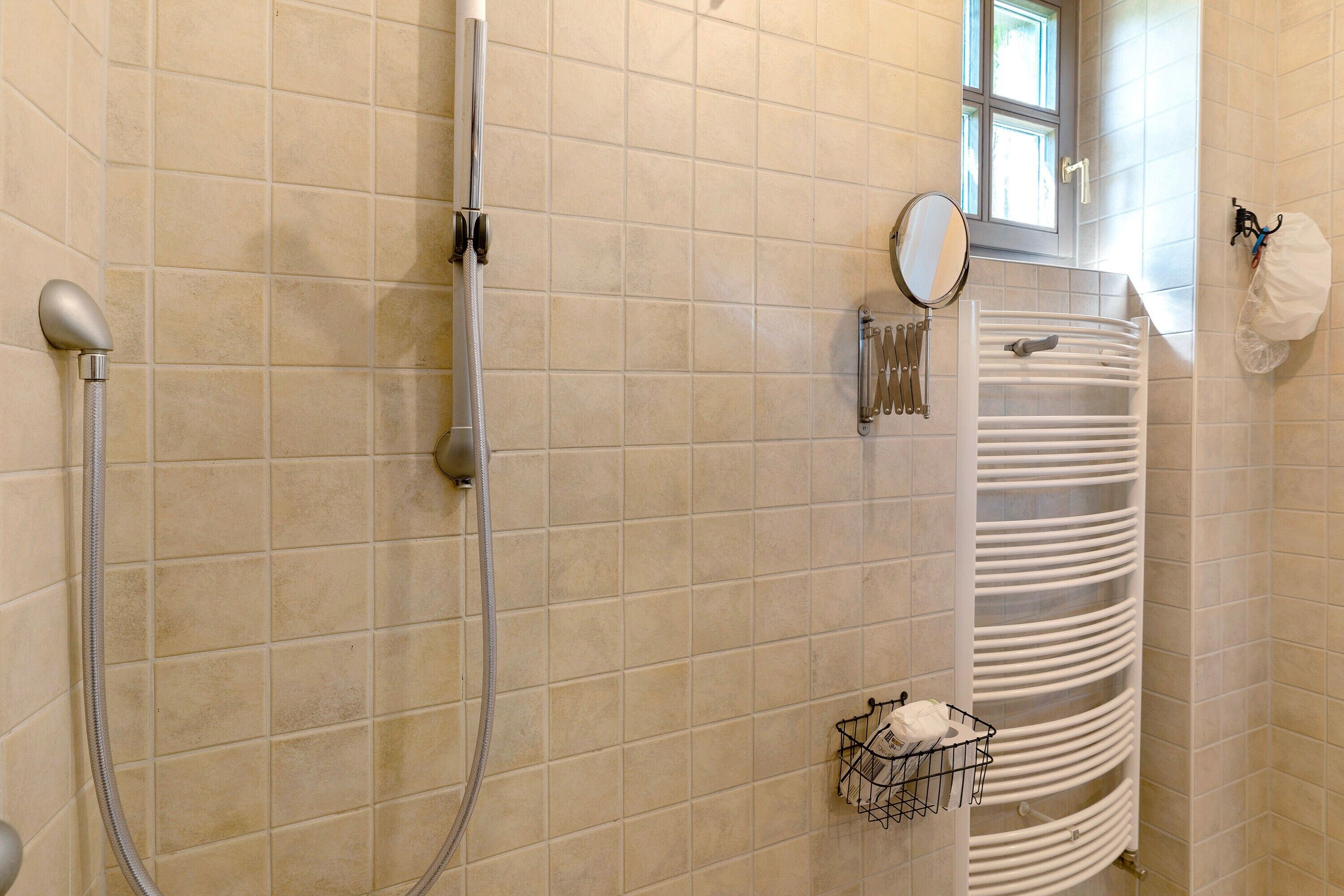 A bathroom with beige tiles, a shower, a towel rack, a mirror, and a window.