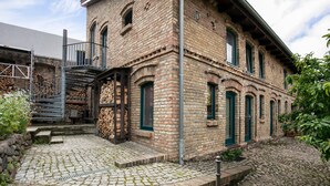 A brick building with a staircase leading to a wooden deck, surrounded by greenery. - Finca 'Gruppenhaus' with Shared Terrace, Communal Garden and Wi-Fi (Groß Kreutz (Havel))