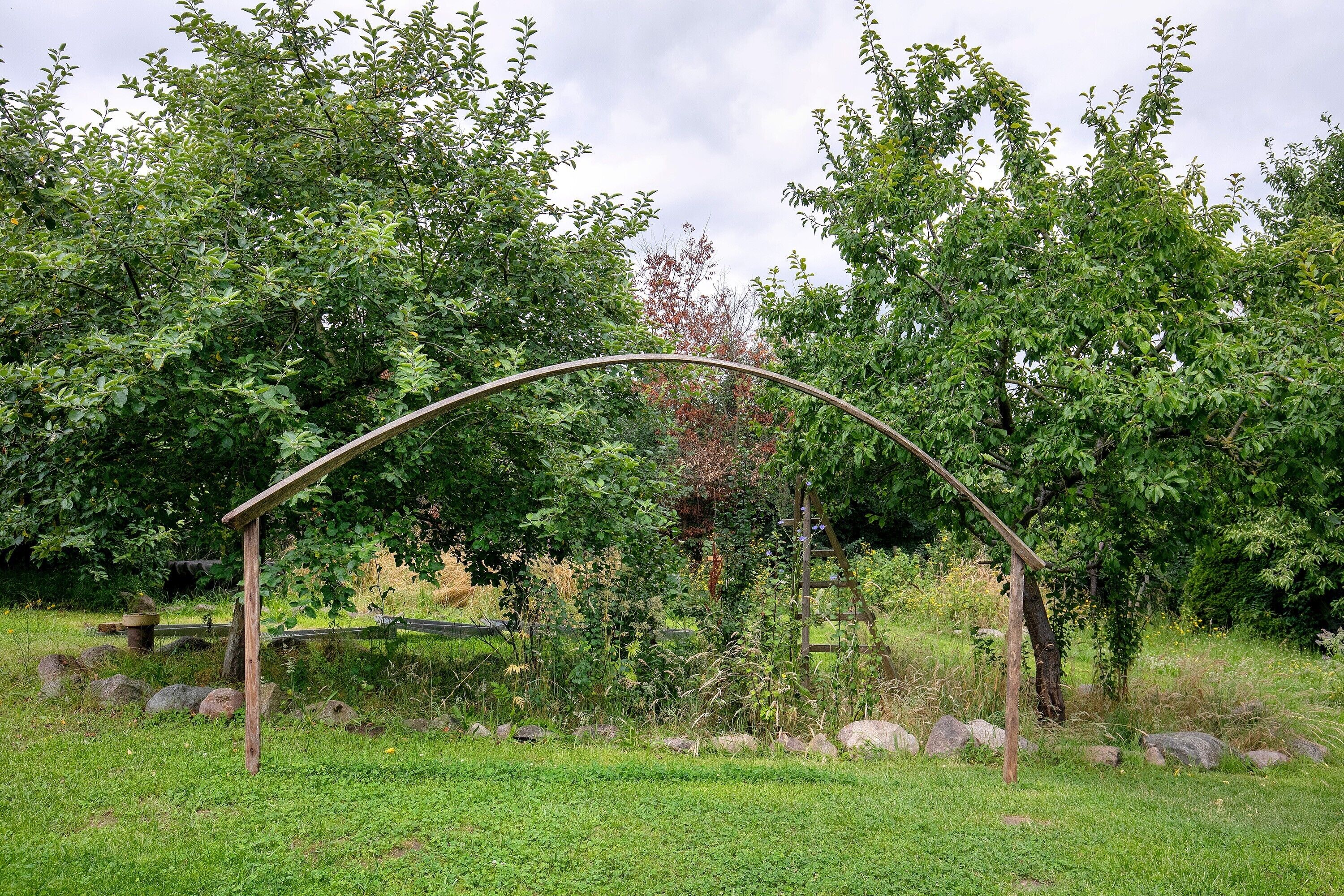A wooden arch structure in a grassy area with trees and rocks.