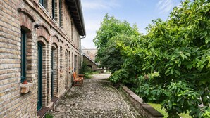A courtyard with a cobblestone path, a brick building with green windows, and a wooden bench. - Finca 'Gruppenhaus' with Shared Terrace, Communal Garden and Wi-Fi (Groß Kreutz (Havel))