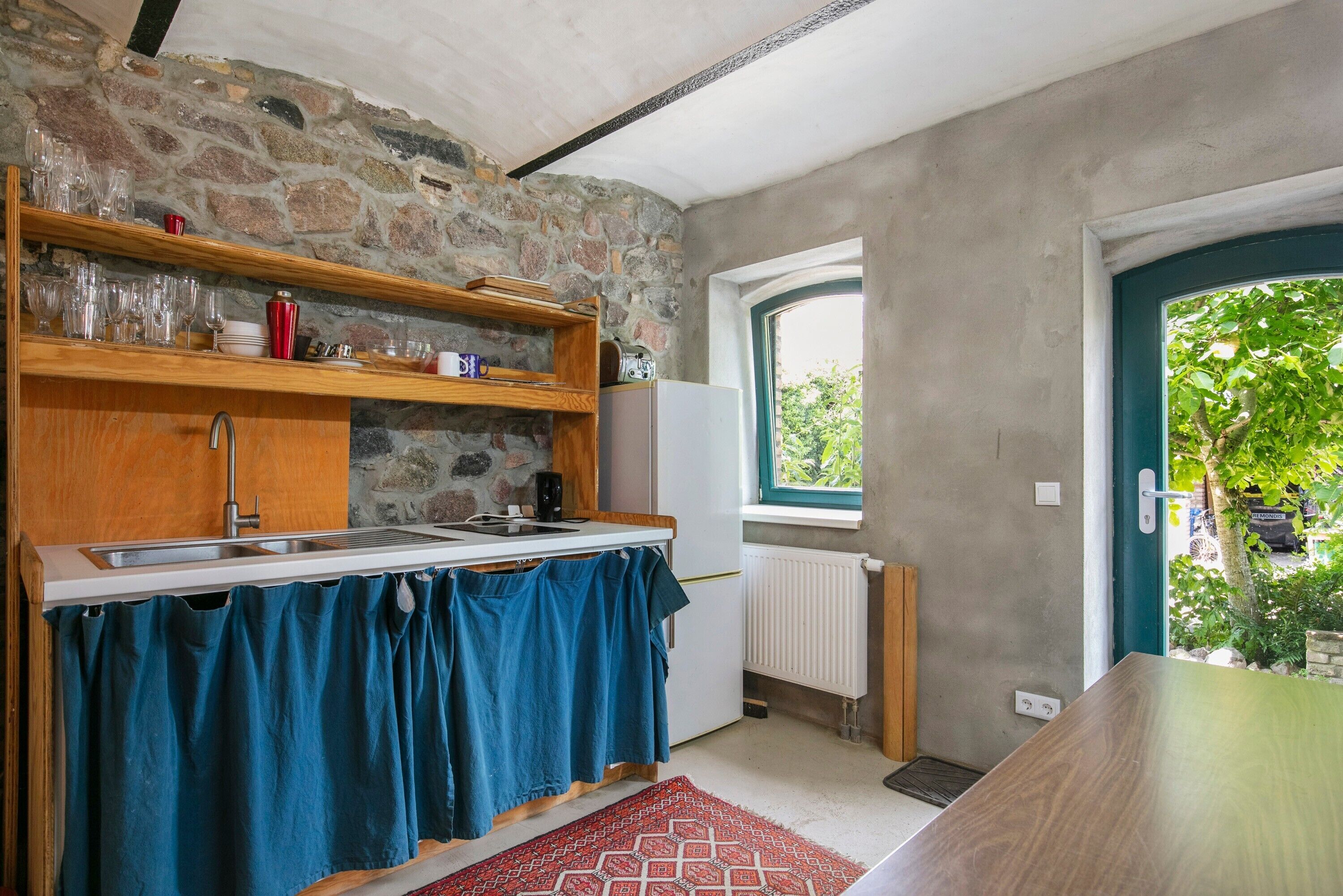 Eine Küche mit Steinwand, Holzböden, einem blauen Vorhang, einem weißen Kühlschrank und einem Fenster mit Blick ins Grüne.
