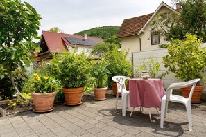 A patio with potted plants, a table with a red and white checkered tablecloth, and white plastic chairs.