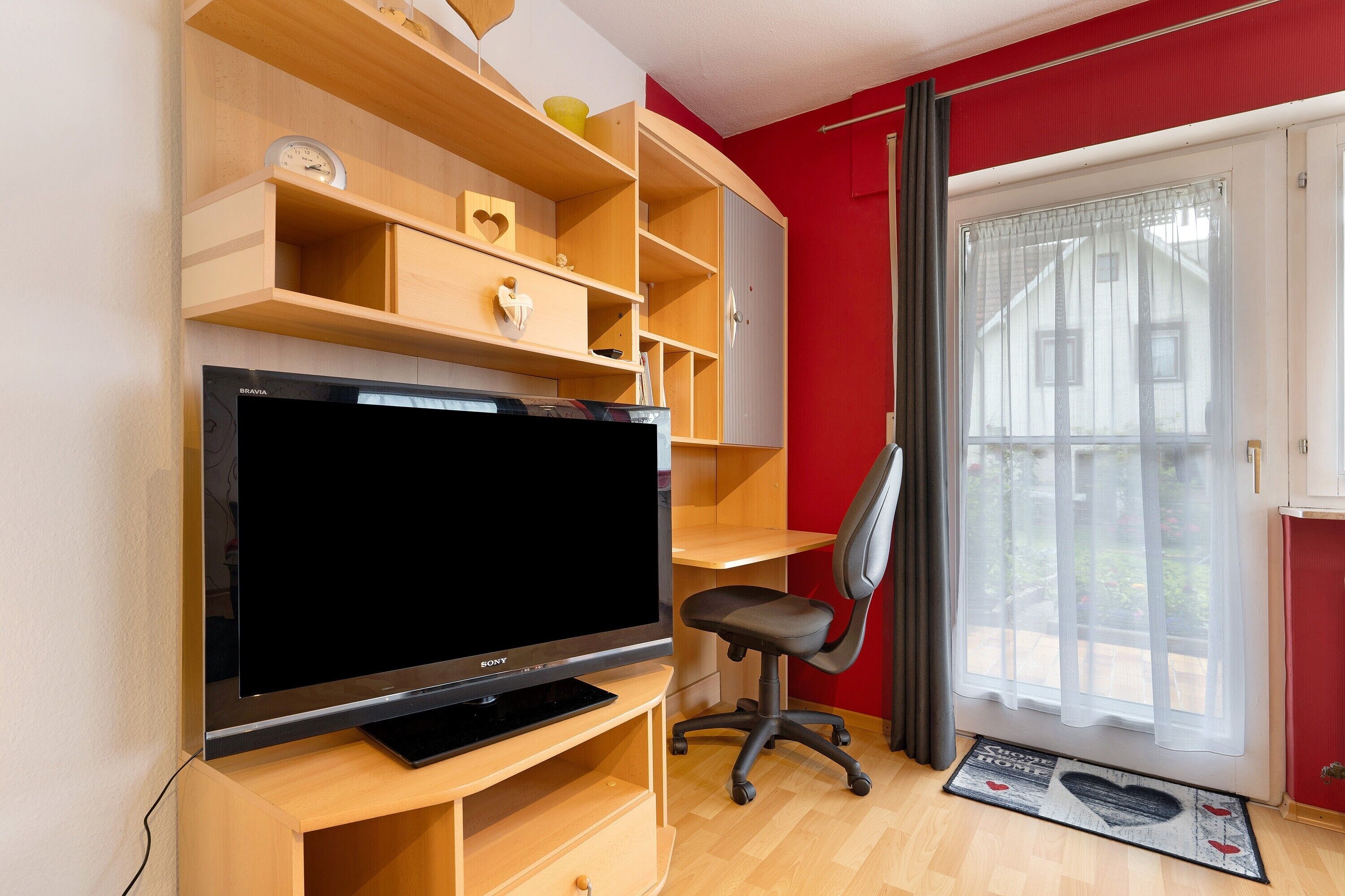 A room with a TV on a wooden stand, a desk with a chair, and a window with curtains.