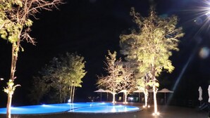 Un espace piscine nocturne agrémenté d’arbres et de parasols illuminés.