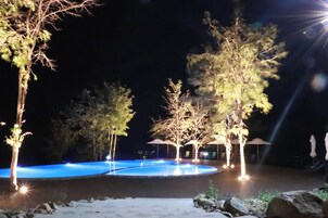 Zona de piscina iluminada de noche, con árboles y sombrillas.