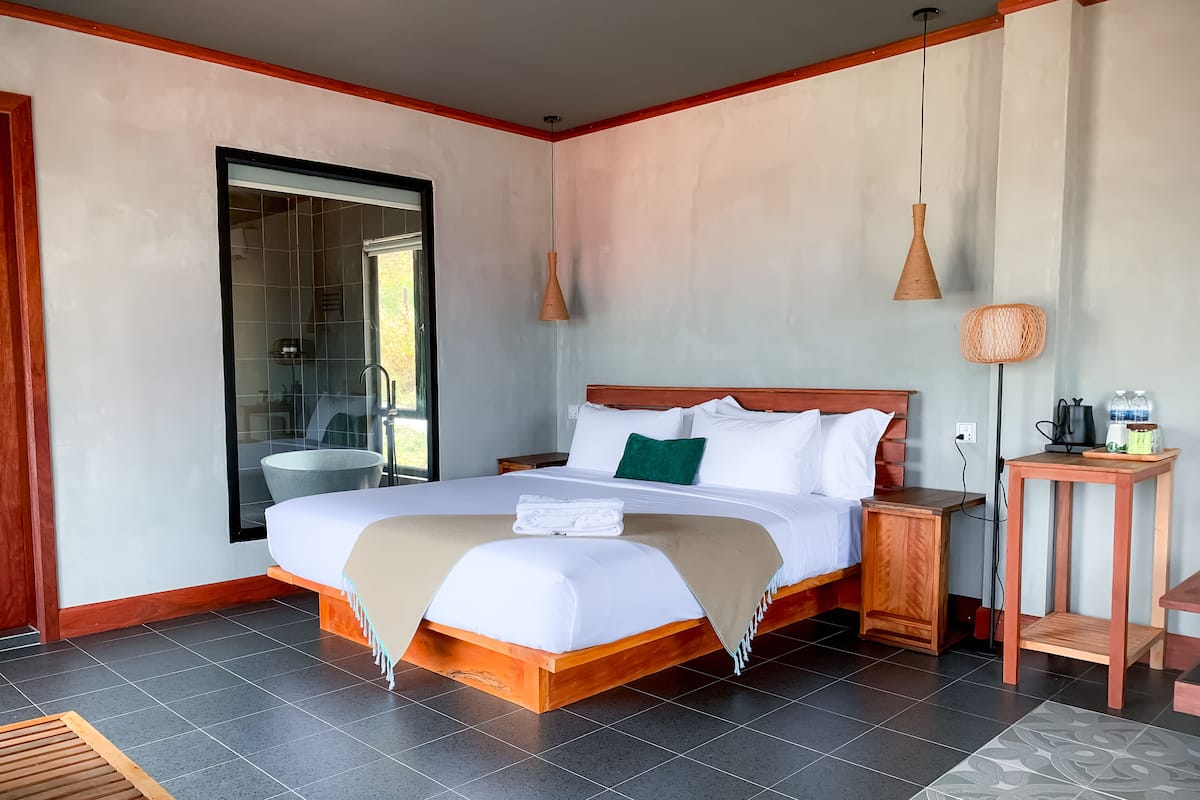 Un dormitorio espacioso con una cama grande, una mesita de noche de madera, una mesita auxiliar y vistas al baño a través de una puerta de vidrio.