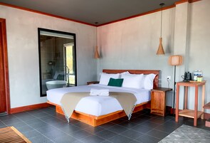 Un dormitorio espacioso con una cama grande, una mesita de noche de madera, una mesita auxiliar y vistas al baño a través de una puerta de vidrio.