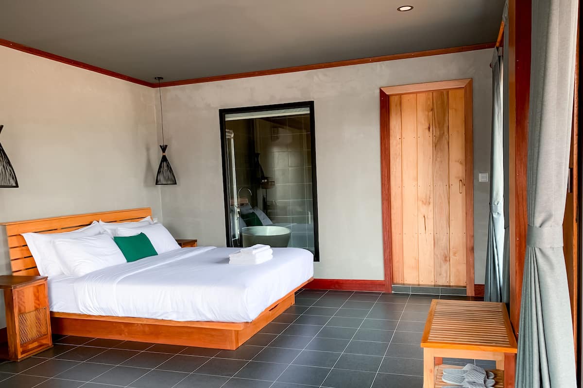 Un dormitorio con cama de madera, una mesita, un armario y un baño visible a través de una puerta de cristal.