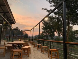 Un espacio de comedor al aire libre con mesas y bancos de madera, rodeado de barandillas de cristal y árboles.