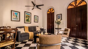 A living room with a checkered floor, a sofa, a coffee table, and a chair. There are framed pictures on the wall, a ceiling fan, and a large wooden door with a decorative arch. - Ocean Breeze | Old San Juan | Spanish Style (San Juan)