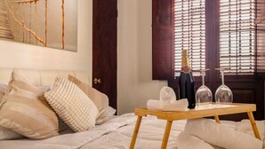 A hotel room with a bed, a wooden tray with a bottle and glasses, and a window with blinds. - Ocean Breeze | Old San Juan | Spanish Style (San Juan)