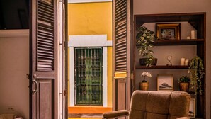A cozy room with a brown armchair, a wooden bookshelf, and a window with shutters. - Ocean Breeze | Old San Juan | Spanish Style (San Juan)