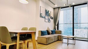 A modern living room with a dining area, a sofa with cushions, a coffee table, and a large window offering a city view. - NVT Housing - Vinhomes Skylake Apartment (Hanoi)