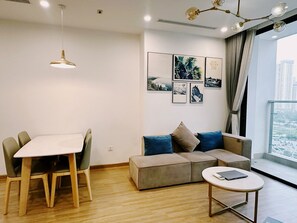 Una sala de estar moderna con una mesa de comedor, un sofá y una pequeña mesa de centro. Hay obras de arte en las paredes y una vista de la ciudad a través de un amplio ventanal.