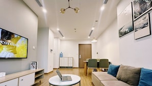 A modern living room with a sofa, a round coffee table with a laptop, a TV on the wall, and a dining area with chairs and a table. - NVT Housing - Vinhomes Skylake Apartment (Hanoi)