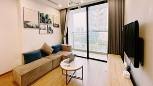 A modern living room with a sofa, a small round table with a laptop, a flat-screen TV mounted on the wall, and large windows offering a city view. - NVT Housing - Vinhomes Skylake Apartment (Hanoi)