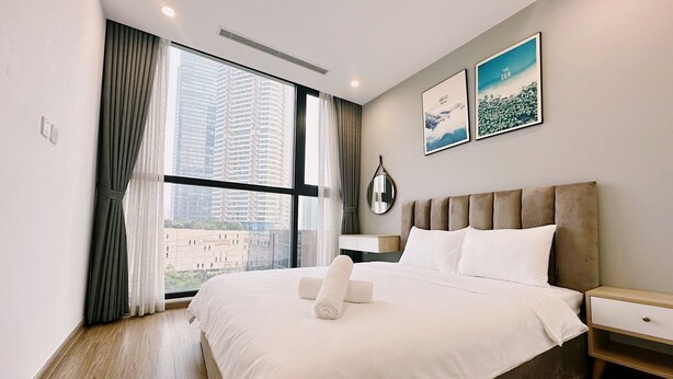 A modern hotel room with a large bed, a bedside table, a mirror, and a view of tall buildings through the window. - NVT Housing - Vinhomes Skylake Apartment (Hanoi)