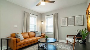 A living room with a brown leather sofa, a round coffee table, a floor lamp, and a side table. There are three windows with curtains, a ceiling fan, and three framed wall art pieces.