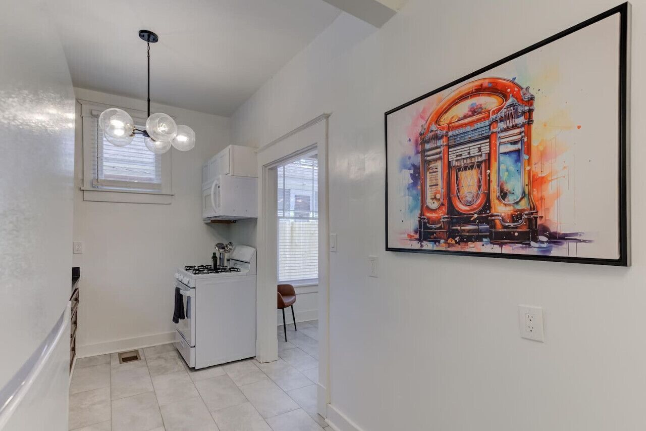 A modern kitchen with white cabinets, a gas stove, and a pendant light. There is a large framed artwork on the wall depicting a colorful building.