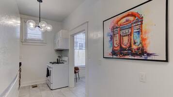 A modern kitchen with white cabinets, a gas stove, and a pendant light. There is a large framed artwork on the wall depicting a colorful building.