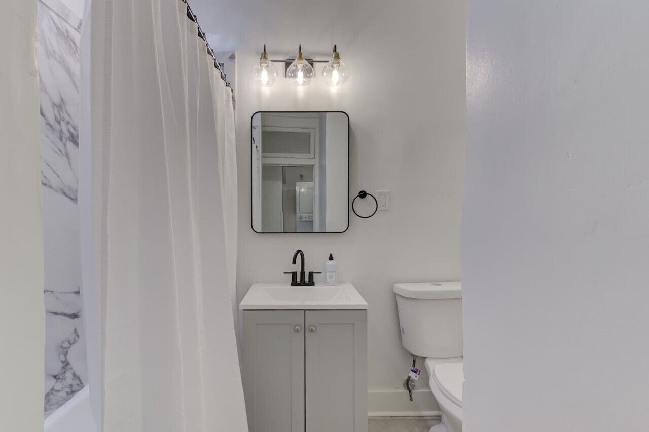 A bathroom with a white sink, a mirror, and a toilet. There are three light fixtures above the mirror.