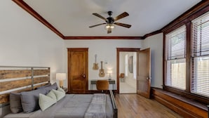 A bedroom with a bed, wooden headboard, a ceiling fan, a guitar, a desk with a chair, and a window with blinds. - The Cooper Suite | Midtown Memphis (Memphis)