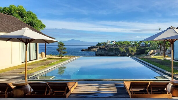 Una piscina infinita con sillones y sombrillas, con vistas a la costa, un promontorio rocoso y vegetación.