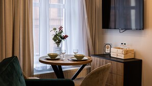 Et hotelværelse med et spisebord dækket til to personer, en vase med blomster, et fjernsyn ophængt på væggen og et træmøbel med en bakke vand på.