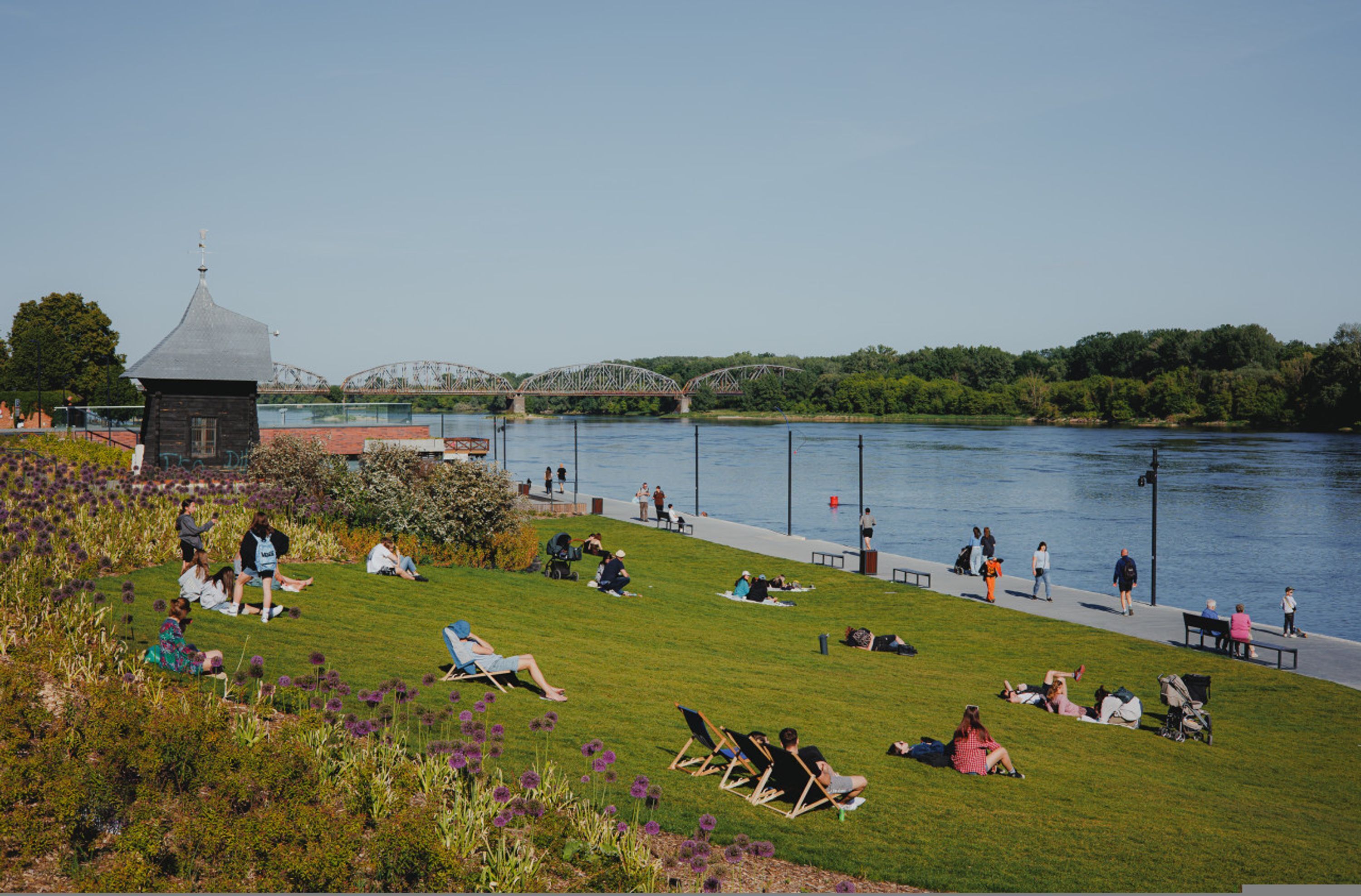 A riverside park with people relaxing on the grass, a bridge in the background, and a small building with a dome roof.