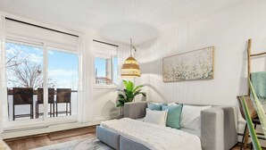 A modern bedroom with a large window, a grey sofa bed, a hanging light fixture, and a potted plant.