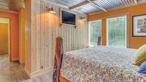 Une chambre avec un mur en bois, un lit recouvert d’une couvre-lit à motifs floraux, un téléviseur fixé au mur et une fenêtre avec des stores.