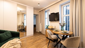 A modern living room with a green sofa, wooden flooring, a dining area with a table and chairs, a TV mounted on the wall, and a large window with curtains. - Estrella 4 Ekskluzywny Pobyt Blisko Restauracji i Kawiarni (Torun)